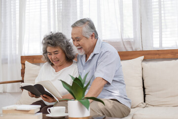 Happy Asian senior couple reading book together on couch in living room, Elderly woman laughing,...