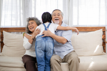 Asian granddaughter running to embracing grandmother and grandfather sitting on couch in the living room. Little girl visiting senior couple at home. Family love and bonding. Healthy living insurance