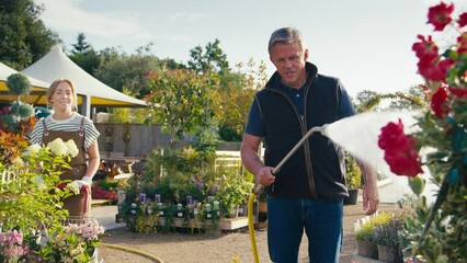 Woman wearing apron pushing barrow of plants working outdoors in garden centre as mature man sprays plants with water from hose - shot in slow motion