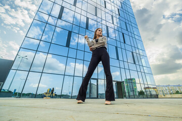 young girl with sunglasses in jacket posing near modern office building with mirrored walls in parking lot with road signs