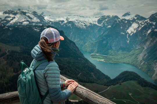 Female hiker with backpack looking at turquoise lake K&ouml;nigssee from above viewpoint of mountain peak Jenner at Berchtesgaden Bavaria, snow-capped mountains in the background.