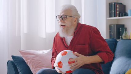 A senior man watches a football soccer game at home, enthusiastically cheering for his favorite team