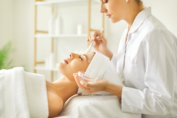 Obraz na płótnie Canvas Woman enjoys professional skin care at spa beauty salon. Side view beautician applying cleansing moisturizing facial mask on face of young woman lying on bed in white towel. Facial treatment concept