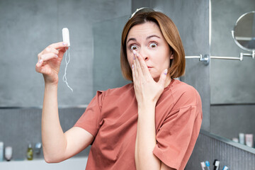 In the bathroom, a woman wears a surprised and dissatisfied expression while holding a tampon, an unexpected inconvenience disrupting her day - obrazy, fototapety, plakaty