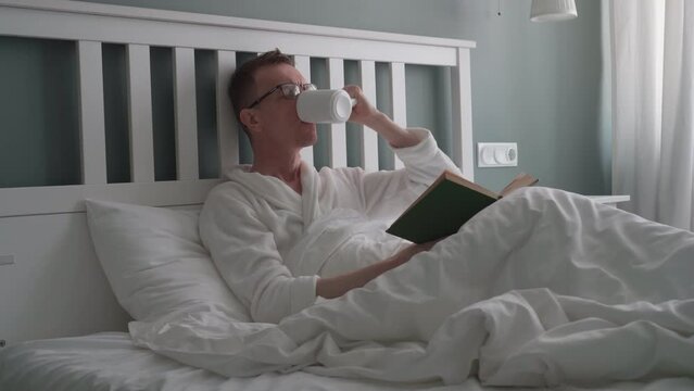Man is lying in bed, reading book and drinking hot drink, tea, milk, soothing decoction from white ceramic cup. Bedroom interior. Natural fabrics and materials. Sleep problems. 4k footag.