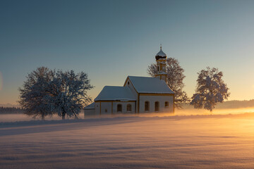 Bavarian church of Raisting with trees and snow and mist during winter and sunset, snow field in the foreground, blue sky day, Bavaria Germany. - 702368624