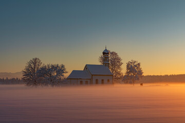 Bavarian church of Raisting with trees and snow and mist during winter and sunset, snow field in the foreground, blue sky day, Bavaria Germany. - 702369022