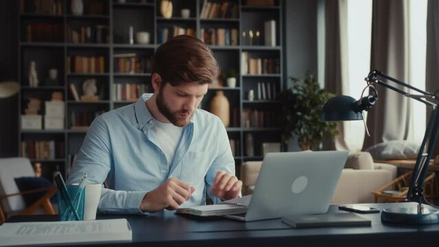 young focused man with beard in blue shirt sits at desktop in home office, works at laptop computer. takes notebook, look through notes thoughtfully, turns pages. types on keyboard, smiles and nods