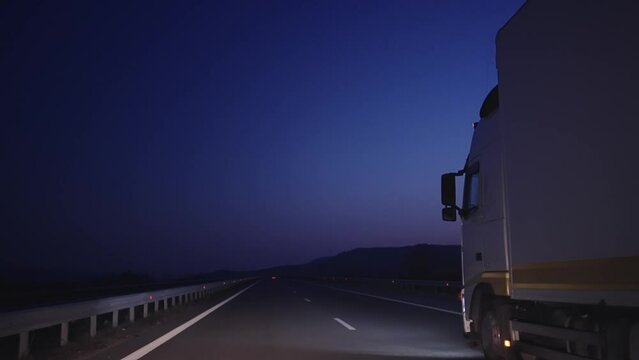 White truck with cargo trailer driving on highway carrying goods at night, insured trucking. Free place on semitrailer for copy space, slow motion cinematic shot