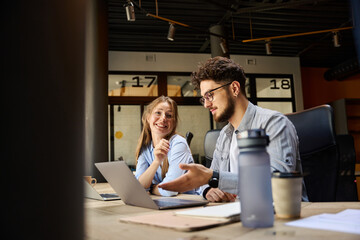 Businesswoman looking at camera while her colleague showing on laptop to her