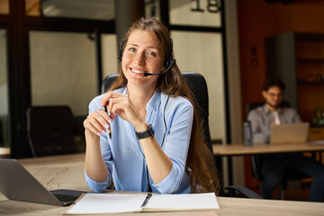 Smiling female call center operator looking at camera near blurred colleague