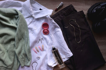 Pastel green cardigan, white shirt, gray trousers, black loafers, various silver jewelry, perfume and make-up products. Outfit concept, top view.