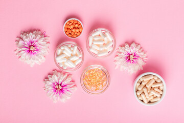 Organic food supplement, minerals and different vitamins such as vitamin D3, C, E in small glass and ceramic jars, pink flowers on pink background top view. Take care of yourself.