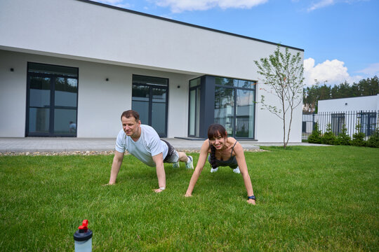 Adult caucasian man and smiling woman doing push ups on green grass in yard