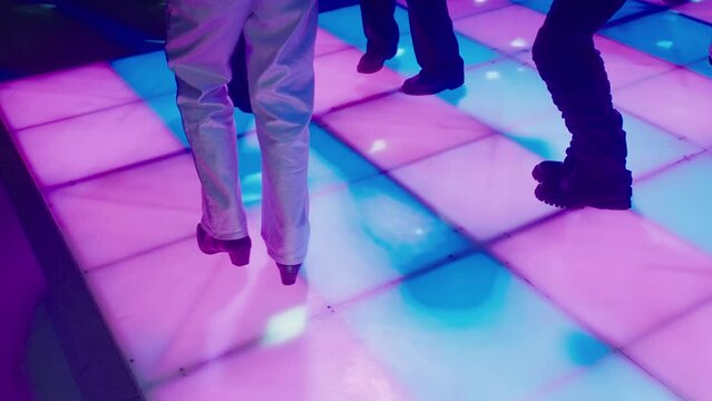 People in bell-bottomed trousers are dancing disco on disco floor glowing with multicolored squares. Vintage disco with dancing legs. Party at a nightclub in the style of 80s. Floor in a nightclub