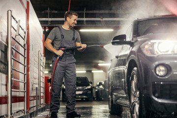 A worker on a car wash station in washing a car under a high pressure water gun.