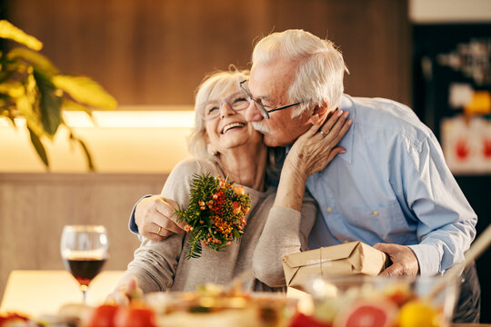 A happy senior couple is hugging and kissing on valentines day and holding gift and bouquet.