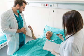 Male and female doctor visiting senior woman in hospital ward writing a report, talking, explaining...