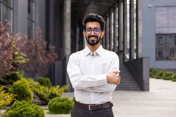 Portrait of a businessman from the outside of the office building, a young successful man smiles and looks at the camera with a crossed hands, an employee in business clothes.