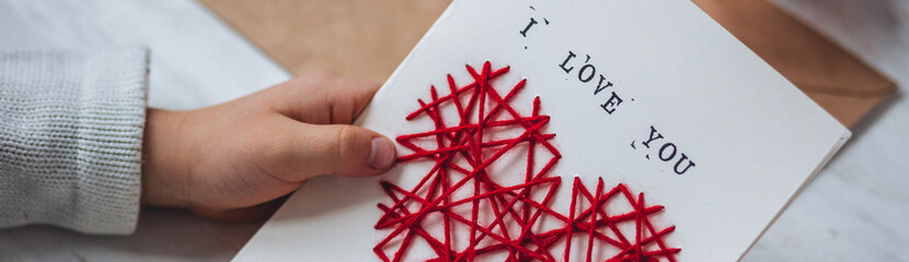 Little kid preparing diy handmade cute post card for Mother's Day with a message I love you mom. Red hear embroidering, printing. Concept of simple presents, family values, hobby, leisure activity