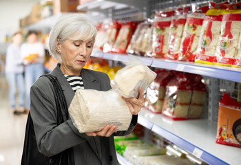 Interested positive senior lady reading label on packages of Asian noodles while shopping in...