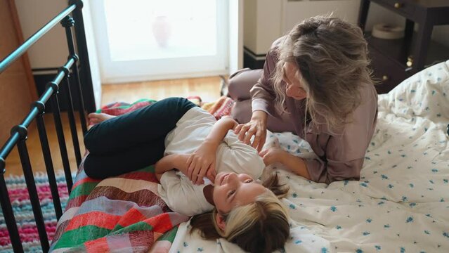 Smiling gray haired young mom tickling her little daughter in pajamas while lying on bed in morning
