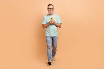 Full size portrait of positive cheerful aged person hold smart phone walking isolated on beige color background