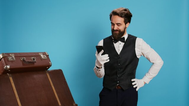 Cheerful employee browses mobile phone, feeling joyful about positive news on online webpages over background. Happy remarkable doorman with white gloves being excited on camera.