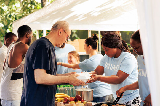 Young volunteers at local center giving complimentary nourishment to diverse group of homeless people benefiting from charitable food campaign. Charity workers share free meals to the hungry and poor.