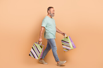 Full size profile portrait of positive aged person hold mall bags walk empty space isolated on...