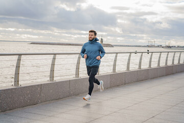 Man in blue jacket enjoys a refreshing run along a waterfront promenade under a cloudy sky.