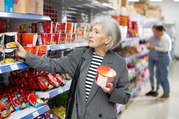 Senior woman in supermarket buys instant fast food in bowl. Mature female buyer reads information...