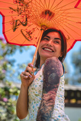 Stunning beauty and japanese style: brunette girl with tattoos, asian dress, and red paper umbrella, delighting in a sunny day with blue sky in a japanese garden