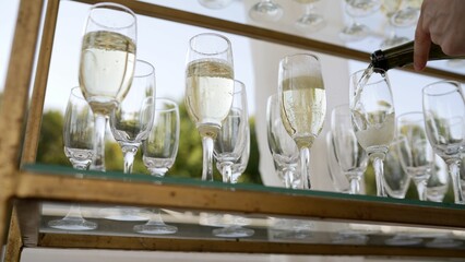 A bottle of sparkling champagne and glasses are poured into a glass at an outdoor wedding event....