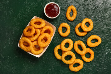 Paper box with fried breaded onion rings and ketchup on green background
