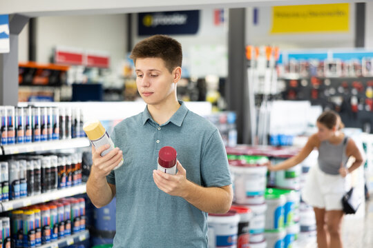 Guy young man chooses picks up buys aerosol paint varnish solvent in construction store. Visitor carefully studies information on product label and buys paint and varnish materials