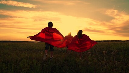 Child imagine being comic book heroes running across meadow in red capes. Children in red capes run across field pretending to be superheroes. Kids actively spend evening dressed as superheroes, dream - obrazy, fototapety, plakaty