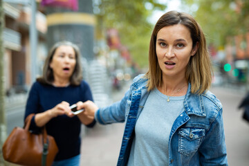 Young woman thief trying to snatch smartphone from hands of elderly passerby on city street and run...