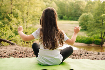 Little athletic girl sitting on sporty rug and practicing yoga in front of river. Active kid meditating in nature.