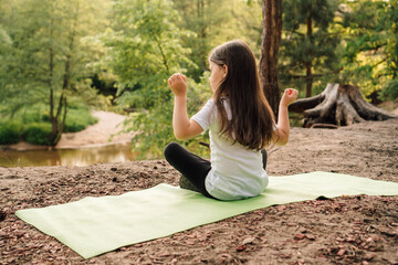 Calm female child sitting on mat in yoga pose with raised arms. Little girl closed eyes and training in front of river.
