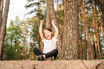 Delightful girl sitting on ground among trees with closed eyes and raised arms. Happy child enjoying activity outdoor.