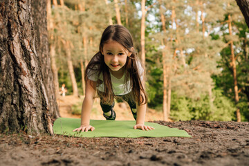 Loveable girl with flowing hair doing plank exercise on green mat outdoor. Junior female athlete working out in nature.