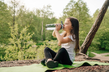 Little girl with dark long hair sitting on green rug in nature and drinking water from bottle, resting after workout.