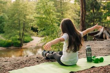 Female kid with arms wide open sitting on mat on sandy hill in front of river with bottle of water and dumbbells near.
