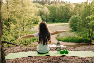 Brunette little girl with arms wide open sitting on green rug on sandy ground. Dumbbells and bottle lying near athlete.