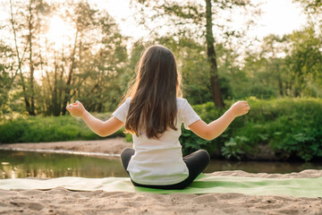 Little girl sit on sporty mat on plage in yoga pose with arms wide open. Kid with dark hair meditating next to lake.