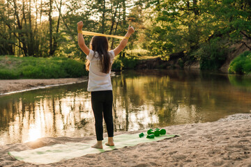 Junior sporty girl raised arms with mini band on wrists standing on mat on sandy ground close to lake, workout outdoor.