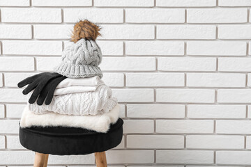 Stack of folded sweaters with hat and gloves on chair against white brick background