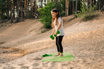 Little lady working out with dumbbells in hands standing on sporty rug outdoor. Junior athlete training on sandy beach.