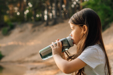 Little girl with dark hair drinking water from bottle sitting outdoor. Active kid resting on sandy...